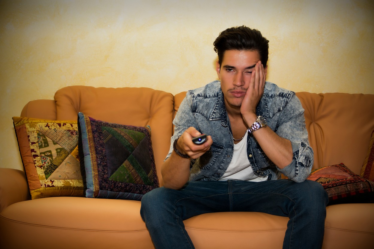 Moški sedi na kavču, v rokah ima daljinski upravljalnik; simbolika sedečega življenjskega sloga, telesne neaktivnosti