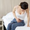 Ženska v kavbojkah in majčki sedi na postelji in se drži za trebuh, simbolični prikaz bolečin v trebuhu, ob menstruaciji, zaradi vnetja ali kakšne druge zdravstvene težave