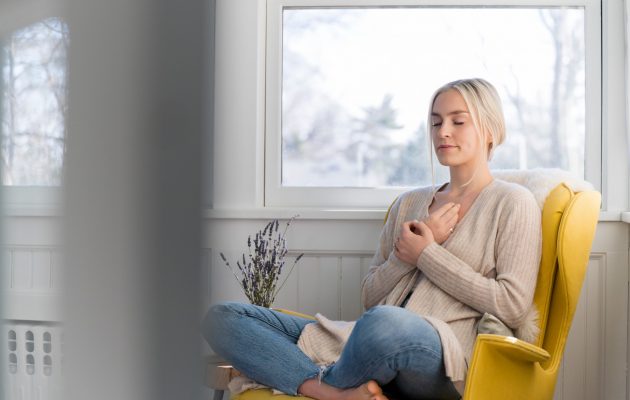 ženska sedi na kavču, roke ima na prsih, oči zaprte - meditira oziroma izvaja vajo čuječnosti