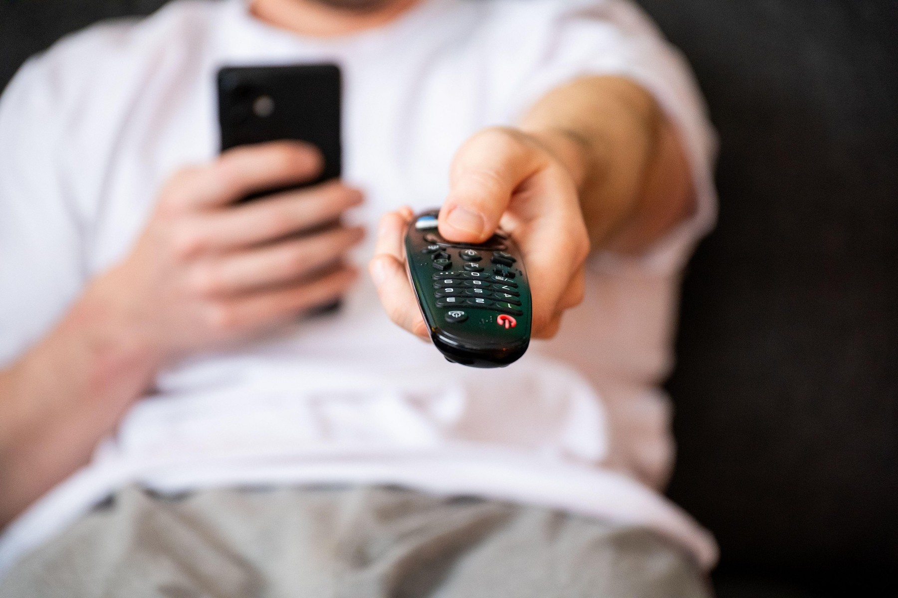 moški drži v rokah telefon in daljinski upravljalnik za televizijo; simbolika sedečega življenjskega sloga, telesne neaktivnosti