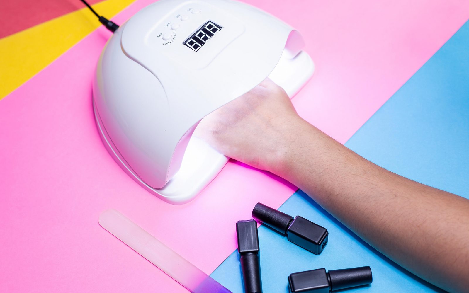 Fotografija prikazuje proces sušenja laka za nohte z uporabo UV/LED lučke. Roka osebe je vstavljena pod belo lučko, ki je postavljena na živahno barvito ozadje, sestavljeno iz roza, rumenih in modrih tonov. Na sliki so vidni tudi trije črni laki za nohte in pilica za nohte. Slika izžareva sodobnost in estetiko ter je primerna za vsebino, povezano z lepotno industrijo, nego nohtov, manikuro, saloni za nohte in domačo nego nohtov.