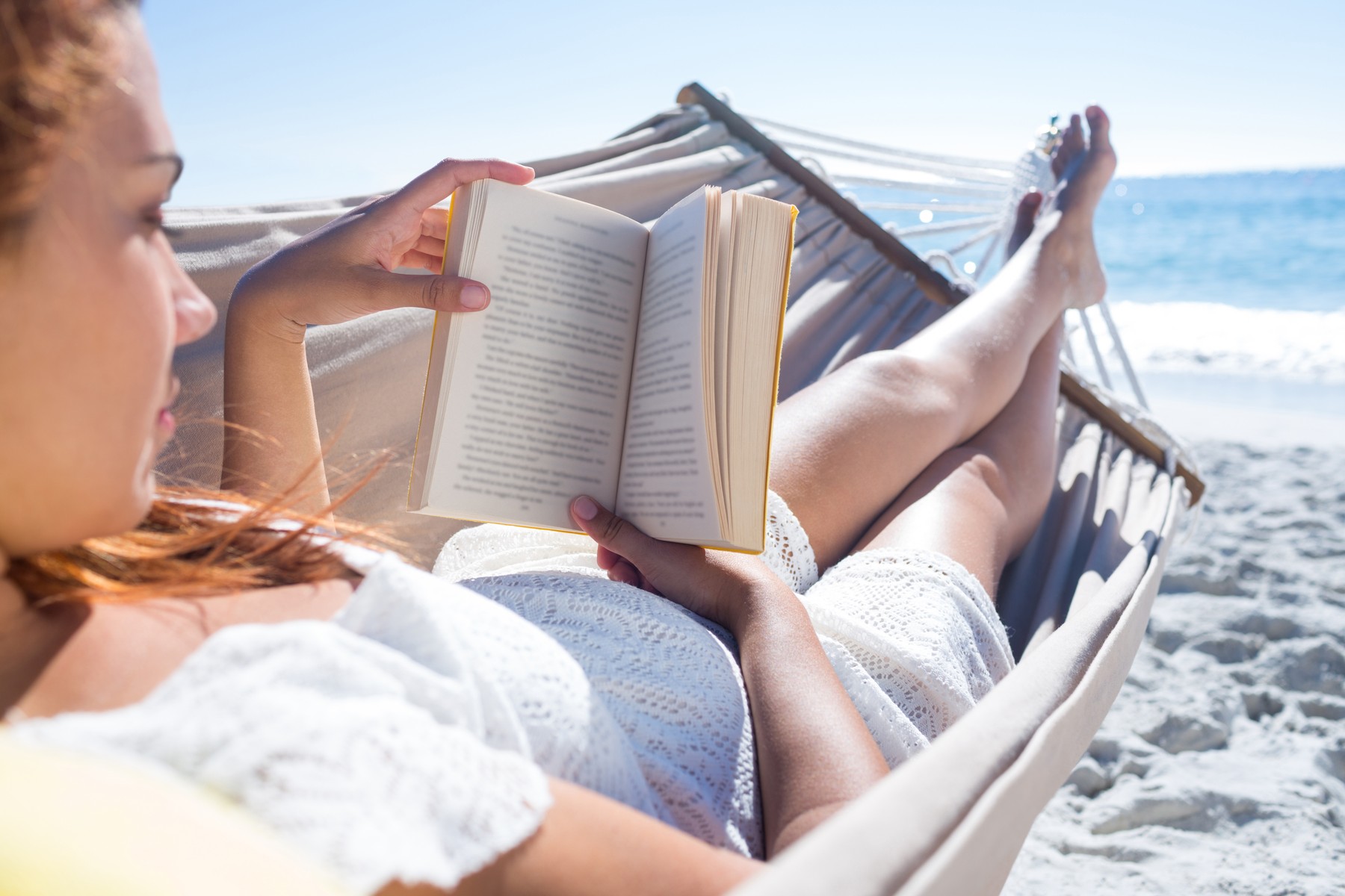 Ženska v beli čipkasti obleki leži v viseči mreži na plaži in bere knjigo. V ozadju je morje in modro nebo. Fotografija izraža sproščenost in mir na sončnem dnevu.