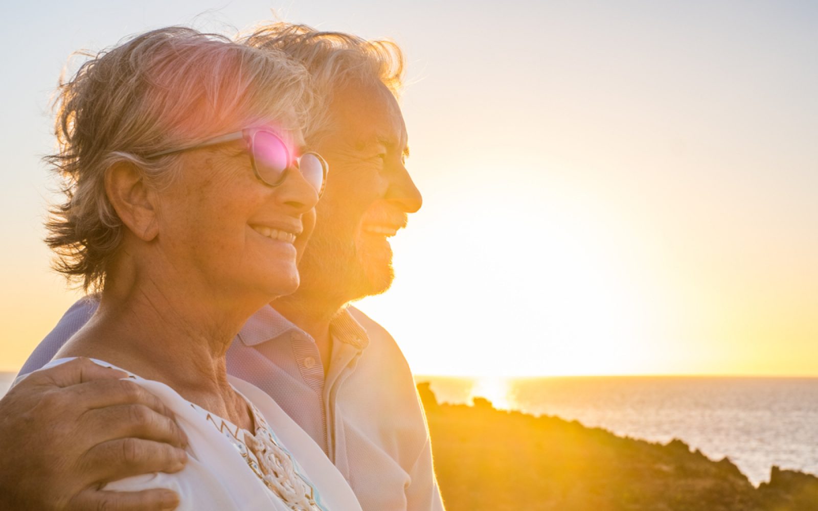 Starejši par uživa ob sončnem zahodu na plaži, nasmejani in objemajoči se, izražajoč srečo, ljubezen in sprostitev. V ozadju se razprostira morje in topla svetloba poletnega večera, kar ustvarja romantičen in miren prizor. Par nosi sproščena poletna oblačila, eden izmed njiju ima na sebi sončna očala, ki odsevajo sončne žarke. Ta slika ponazarja srečne trenutke in uživanje prostega časa v naravi med počitnicami.