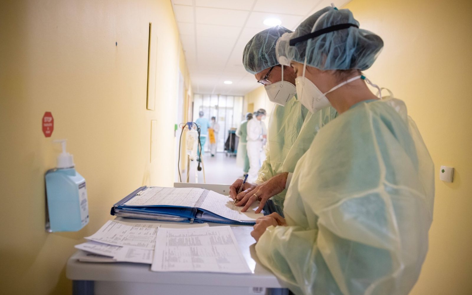 Na sliki so zdravstveni delavci, ki nosijo zaščitno opremo, vključno z maskami, rokavicami in zaščitnimi oblekami, ki delajo v bolnišnici. Dva zdravstvena delavca pišeta zapiske na vozičku, na katerem so tudi različni dokumenti in beležke. V ozadju je bolnišnični hodnik, kjer so še drugi zdravstveni delavci. Na steni je obešena steklenička za razkuževanje rok. Scena prikazuje prizadevanja zdravstvenega osebja pri zagotavljanju oskrbe bolnikom v zahtevnih razmerah.