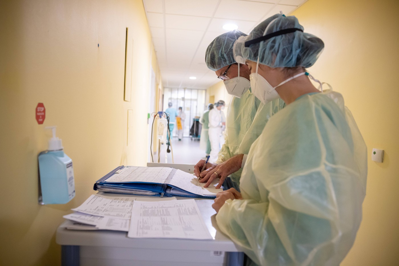 Na sliki so zdravstveni delavci, ki nosijo zaščitno opremo, vključno z maskami, rokavicami in zaščitnimi oblekami, ki delajo v bolnišnici. Dva zdravstvena delavca pišeta zapiske na vozičku, na katerem so tudi različni dokumenti in beležke. V ozadju je bolnišnični hodnik, kjer so še drugi zdravstveni delavci. Na steni je obešena steklenička za razkuževanje rok. Scena prikazuje prizadevanja zdravstvenega osebja pri zagotavljanju oskrbe bolnikom v zahtevnih razmerah.