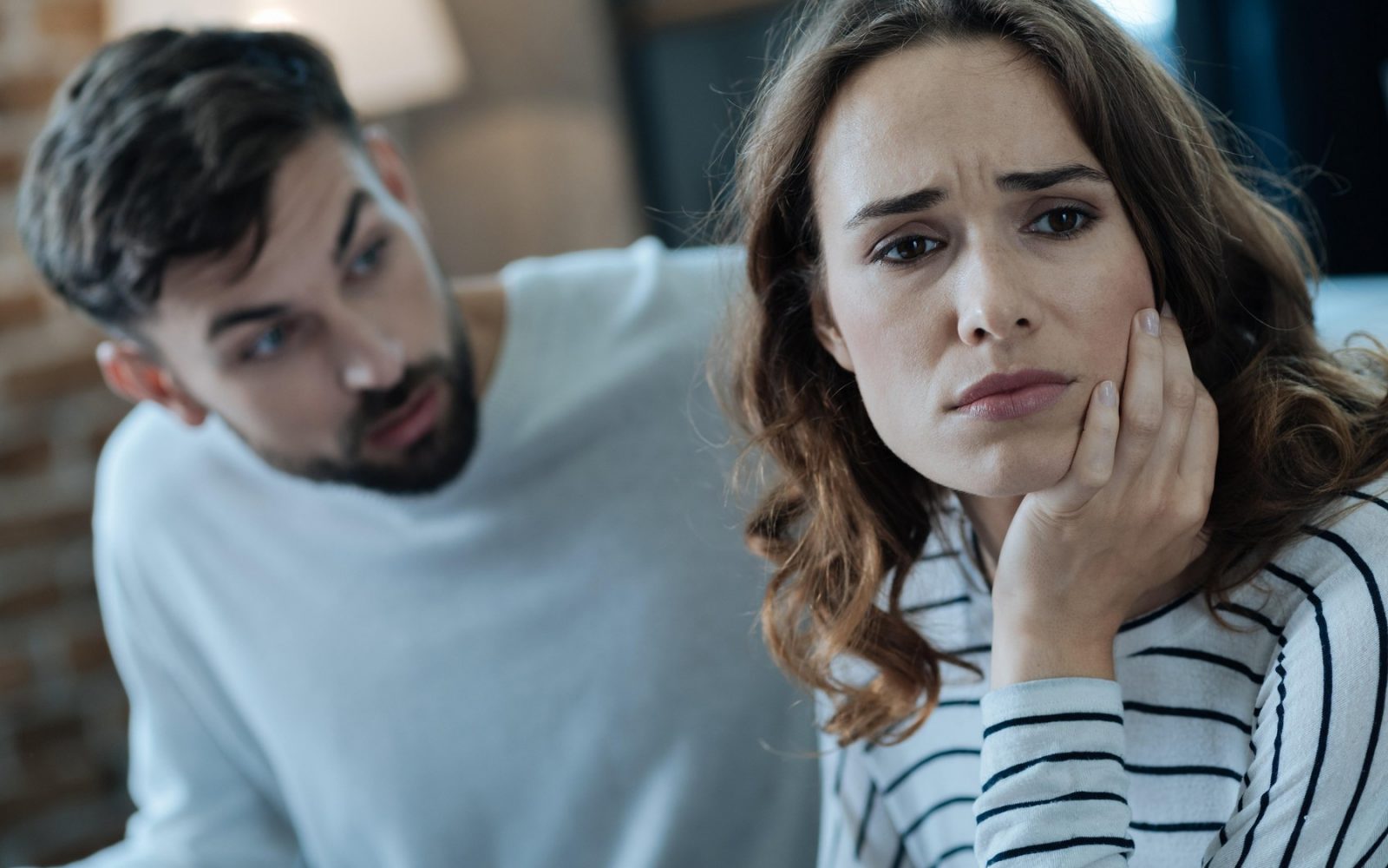 Na sliki je par, ki je v intenzivnem pogovoru. Ženska je videti zaskrbljena ali zamišljena, s podporo glave na roki, medtem ko moški govori z njo. Oba sta oblečena v priložnostna oblačila in sta v notranjem prostoru. Ozadje je nekoliko zamegljeno, vendar se zdi, da je soba udobno opremljena.