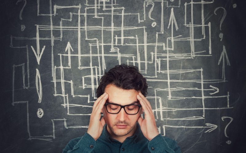 Moški z očali se drži za glavo, na tabli za njim pa je zmeda s puščicami in vprašanji, kar simbolizira težke odločitve ali stres.