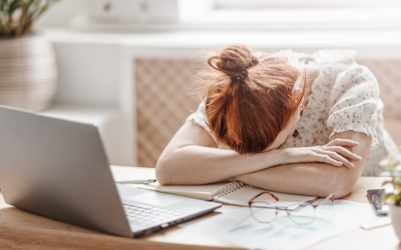 Utrujena ženska, ki sedi za računalnikom, naslonjena na pisalno mizo, kar simbolizira izgorelost in stres pri delu.