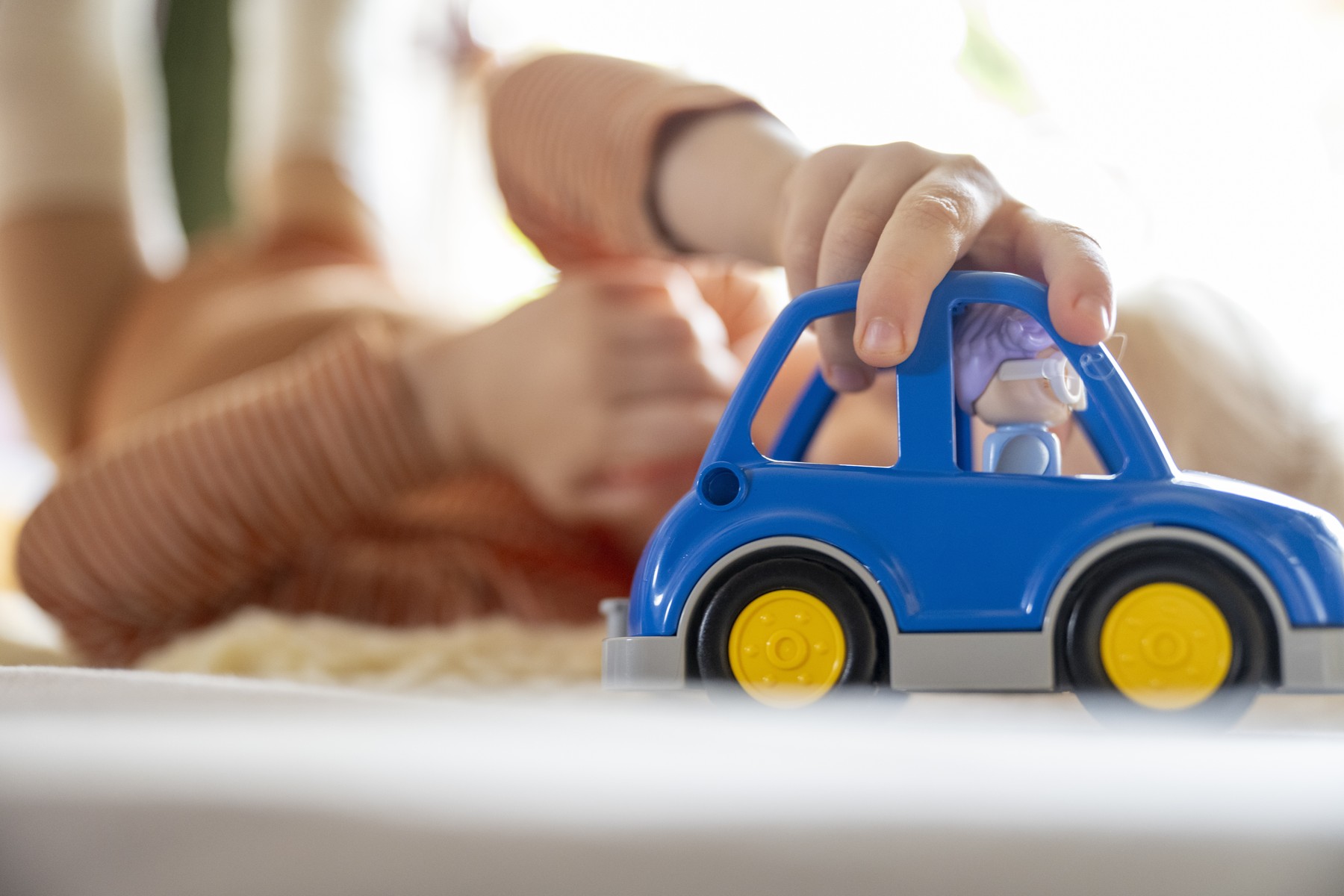 Otrok drži modri avtomobilček z rumenimi kolesi med igro na svetlem ozadju.
