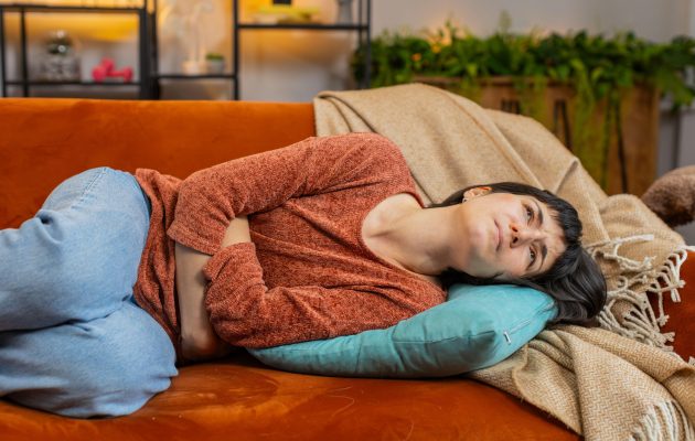 Utrujena ženska leži na kavču in se drži za trebuh, ki jo boli