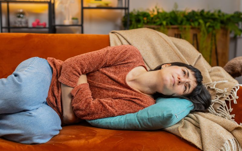 Utrujena ženska leži na kavču in se drži za trebuh, ki jo boli