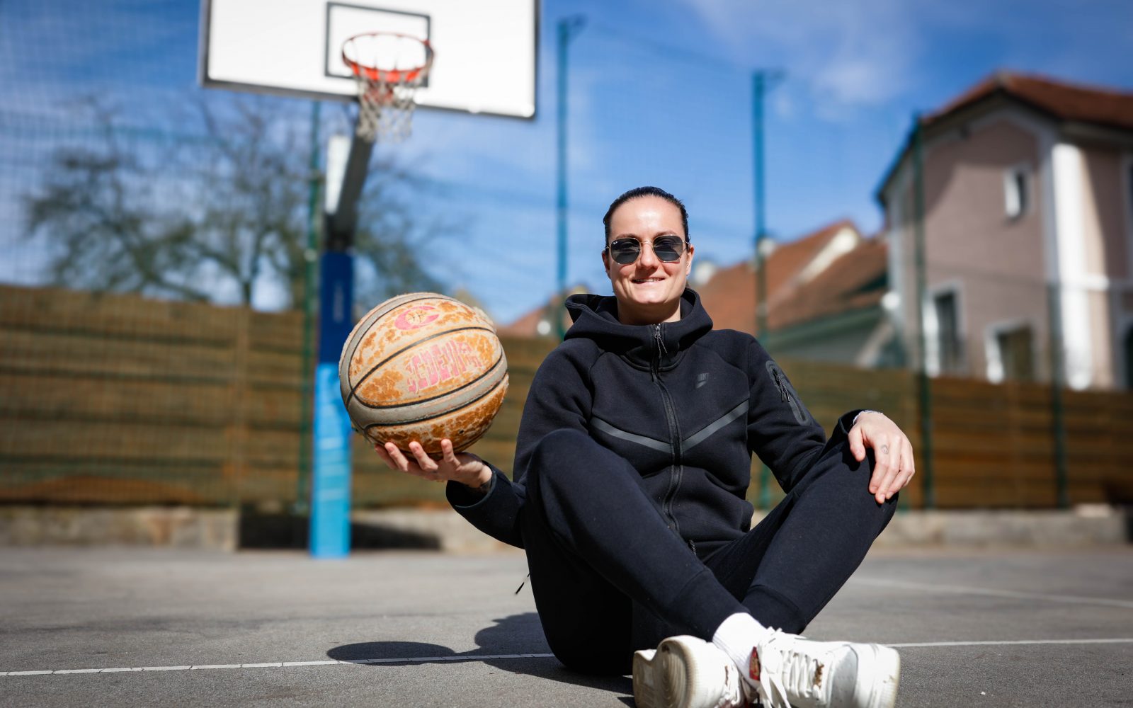 Nika Barič sedi ob košarkarskem igrišču s košarkarsko žogo, ob sončnem dnevu.
