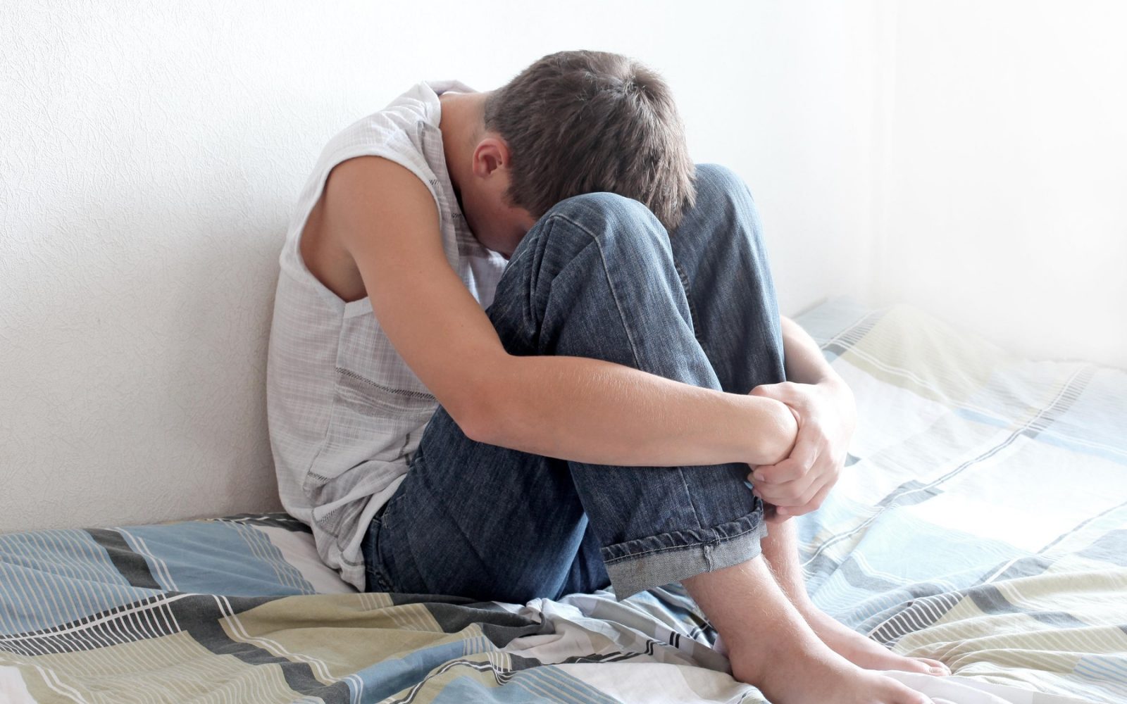 Žalosten najstnik sedi na postelji v domačem okolju, sklonjen in objema svoja kolena.