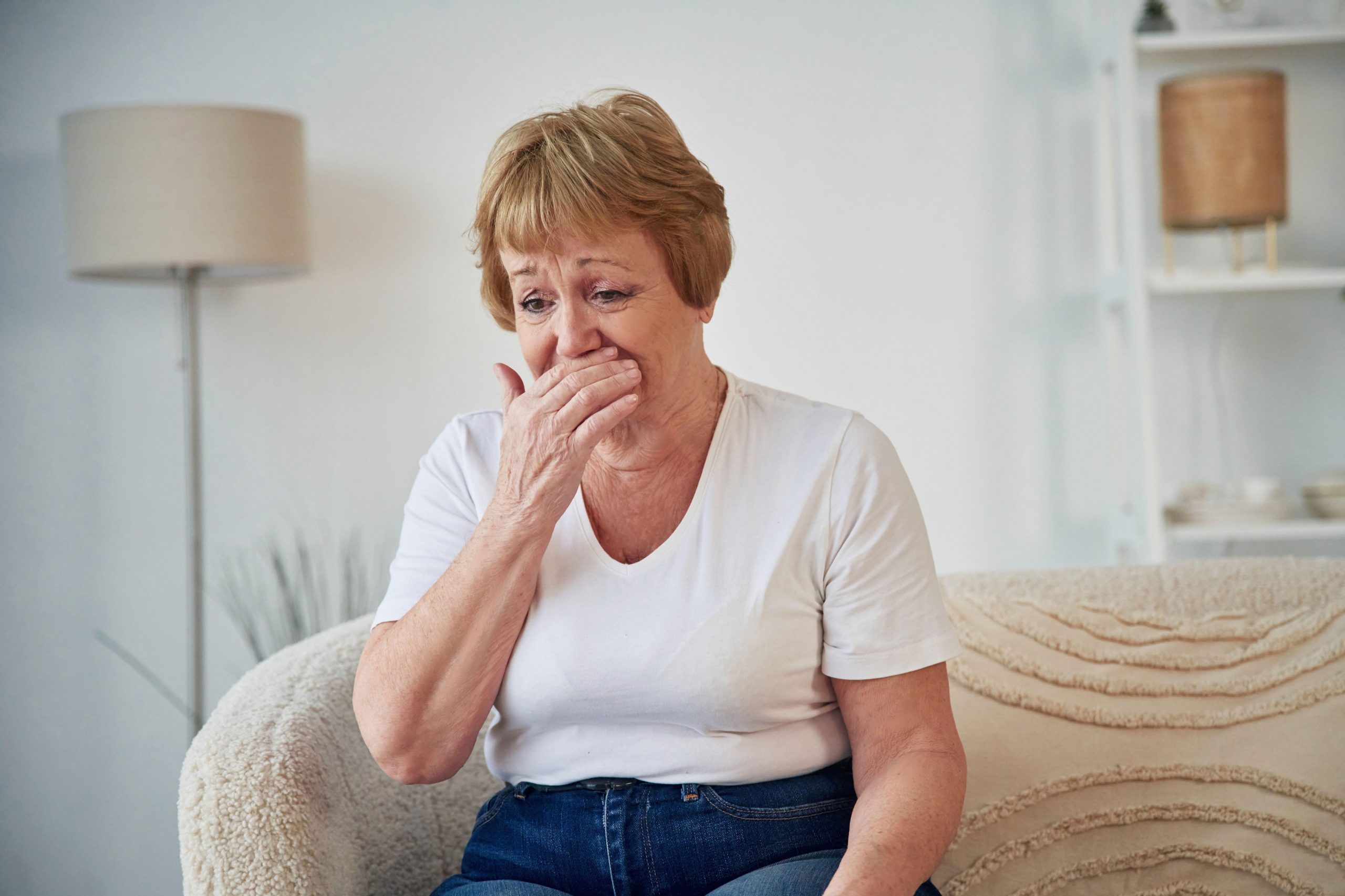 Starejša ženska sedi na kavču in kašlja, kašelj je simptom pljučnega raka
