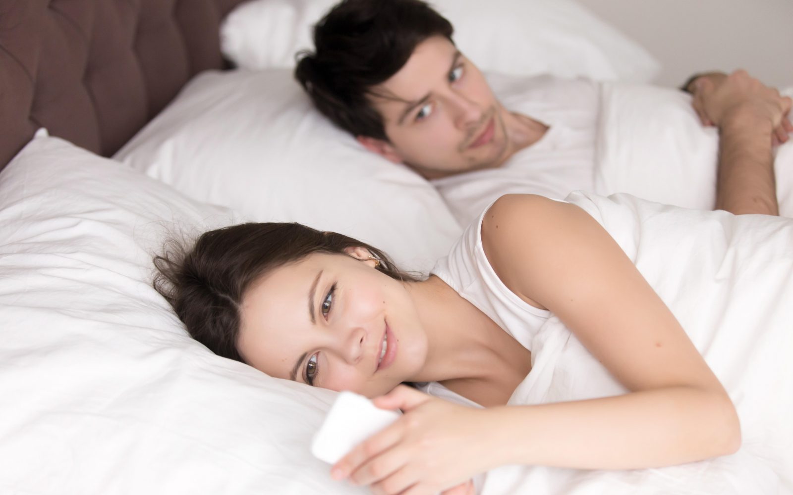 Par ženska in moški ležita v postelji. Ženska gleda v telefon in se smehlja, moški sumičavo opazuje. Mikrovaranje, nezvestoba, pošiljanje sporočil