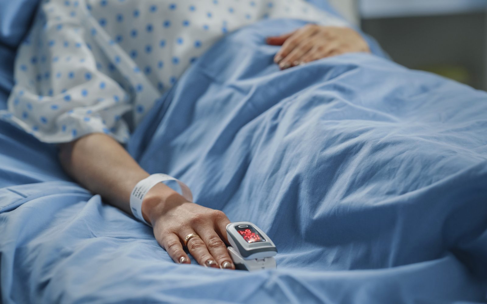 Ženska leži v bolniški postelji, na prstu ima oksimeter