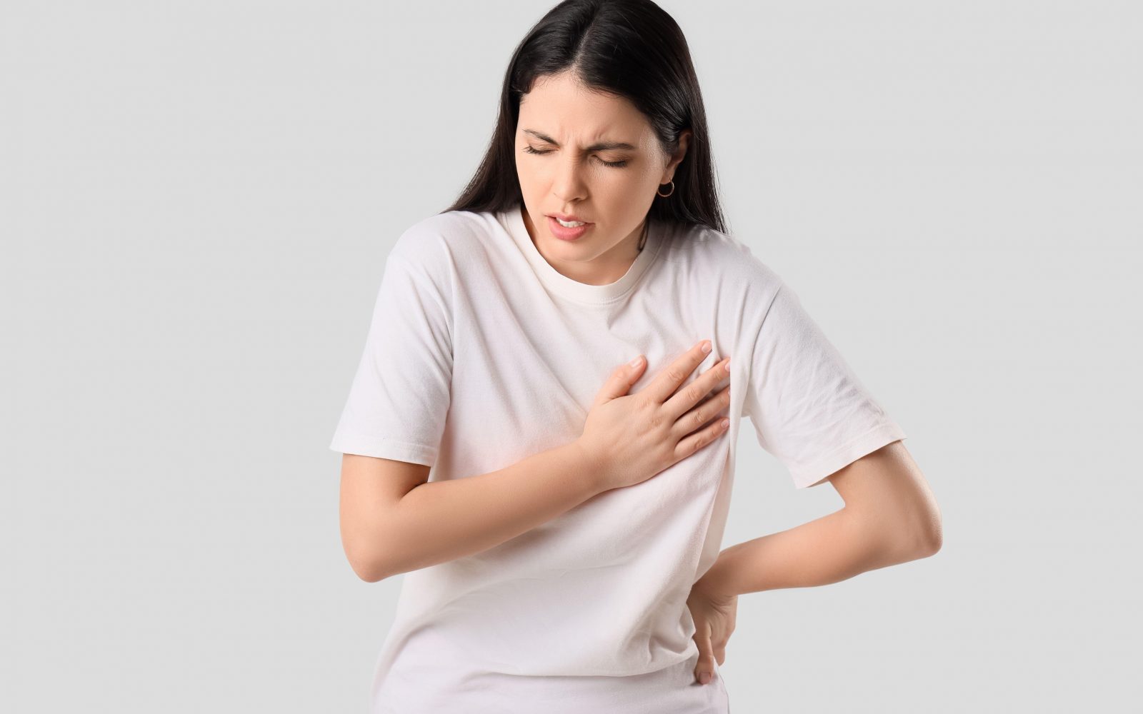 Mlada ženska z bolečinami v prsih na belem ozadju. Lahko gre za infarkt ali težave s srcem, morda pa je kriv tudi napad panike ali hude tesnobe.