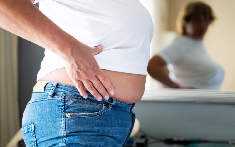 Oseba z odvečnimi kilogrami se kritično opazuje v ogledalu – ponazoritev težav z debelostjo in samopodobo