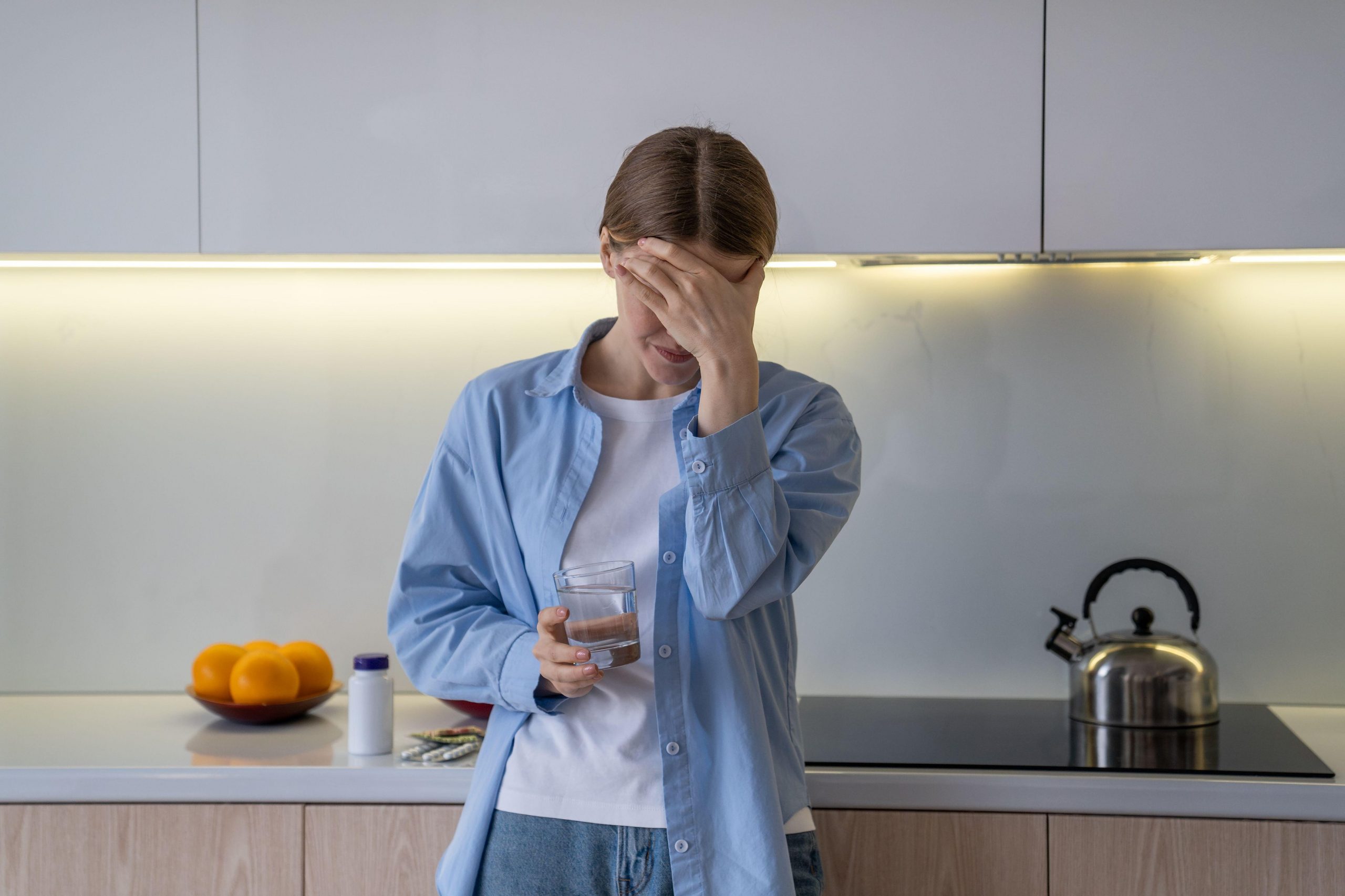 Ženska stoji v kuhinji in se drži za glavo zaradi glavobola, ki je posledica dehidracije. Je v bolečinah in slabe volje, v roki pa drži kozarec vode.