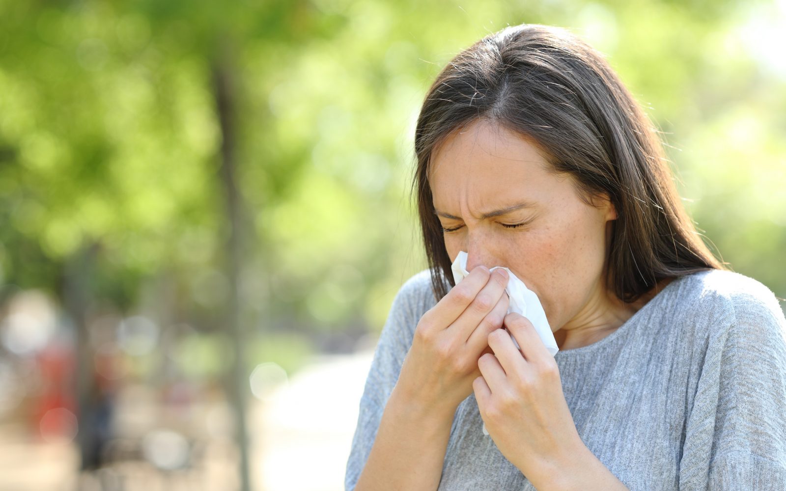 Ženska, ki na prostem kihne v robček. Razlog je morda prehlad, morda pa sezonska alergija na cvetni prah ali dražeči delci v zraku.