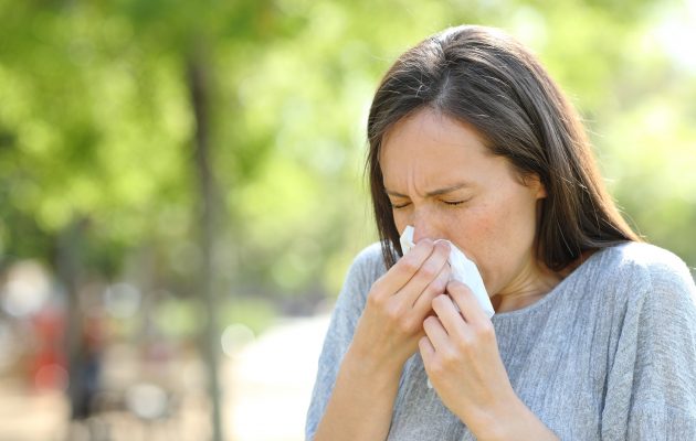 Ženska, ki na prostem kihne v robček. Razlog je morda prehlad, morda pa sezonska alergija na cvetni prah ali dražeči delci v zraku.