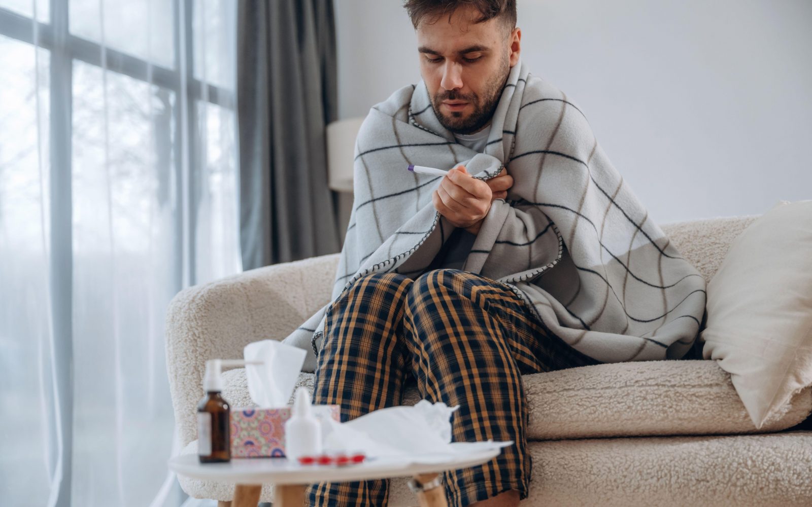 Prehlajen moški, ovit v odejo, sedi na kavču in si s termometrom meri temperaturo, pred njim je miza, na kateri so zdravila za lajšanje simptomov prehlada, kot so sirup, tablete, pršilo za nos in robčki.