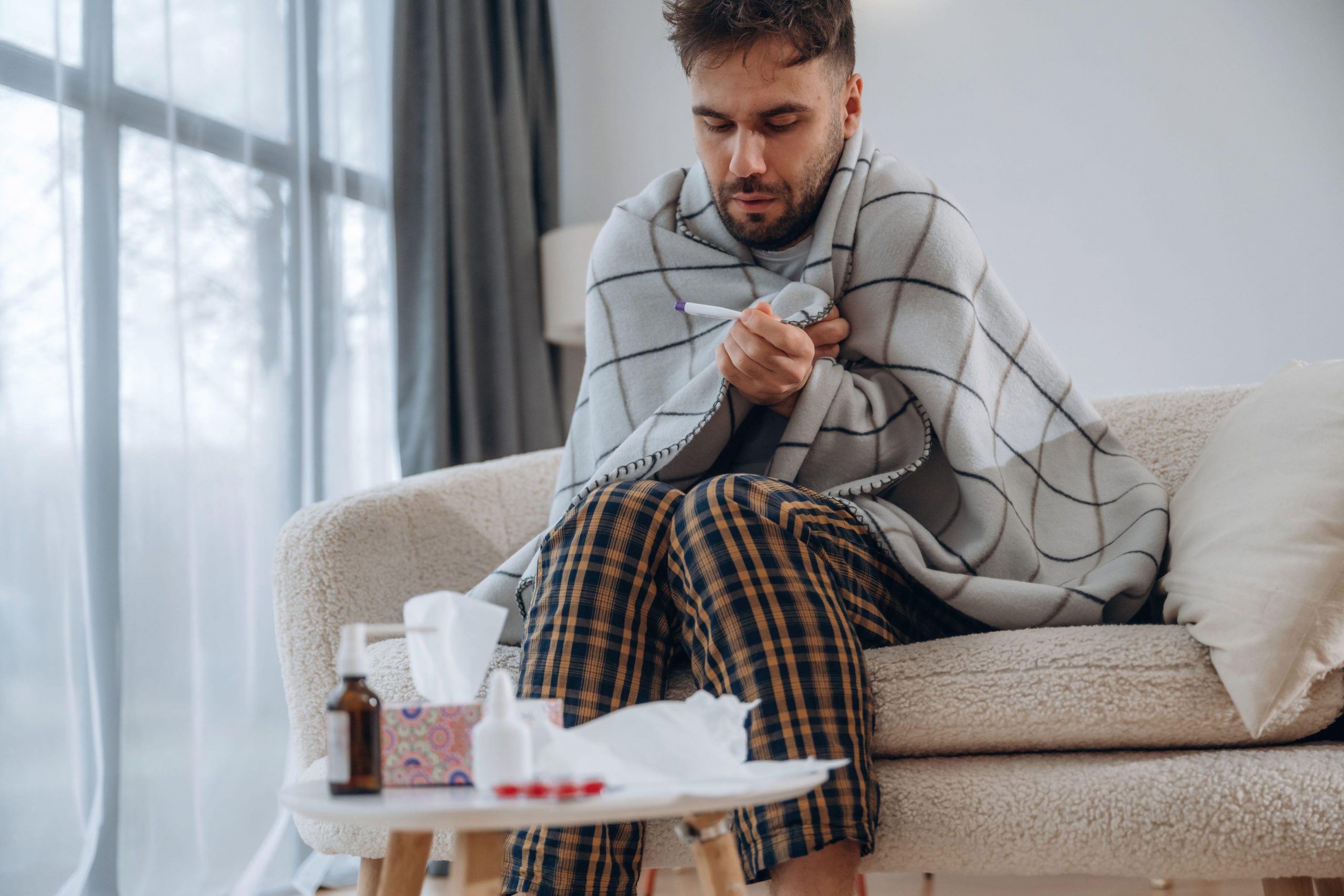 Prehlajen moški, ovit v odejo, sedi na kavču in si s termometrom meri temperaturo, pred njim je miza, na kateri so zdravila za lajšanje simptomov prehlada, kot so sirup, tablete, pršilo za nos in robčki.