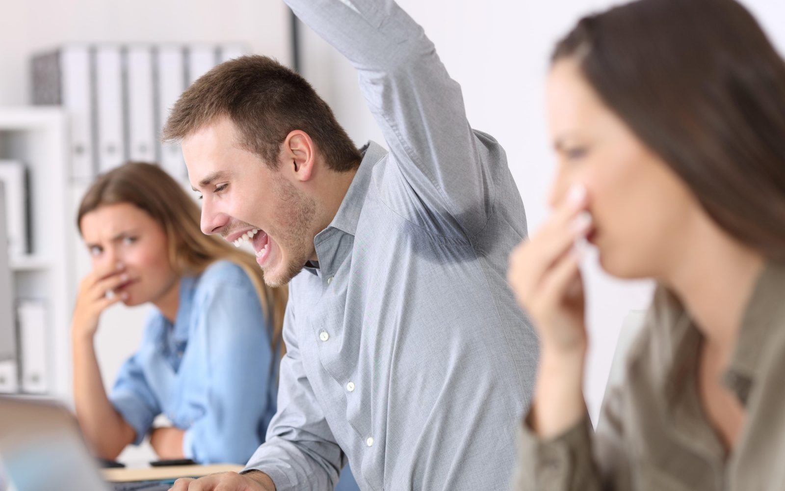 Na fotografiji je moški v sivi srajci, ki navdušeno dvigne roko, a ima pod pazduho velik madež od potu. Dve sodelavki v ozadju si z roko pokrivata nos in kažeta znake nelagodja zaradi neprijetnega vonja. Slika simbolizira problem telesnega vonja v službenem okolju.