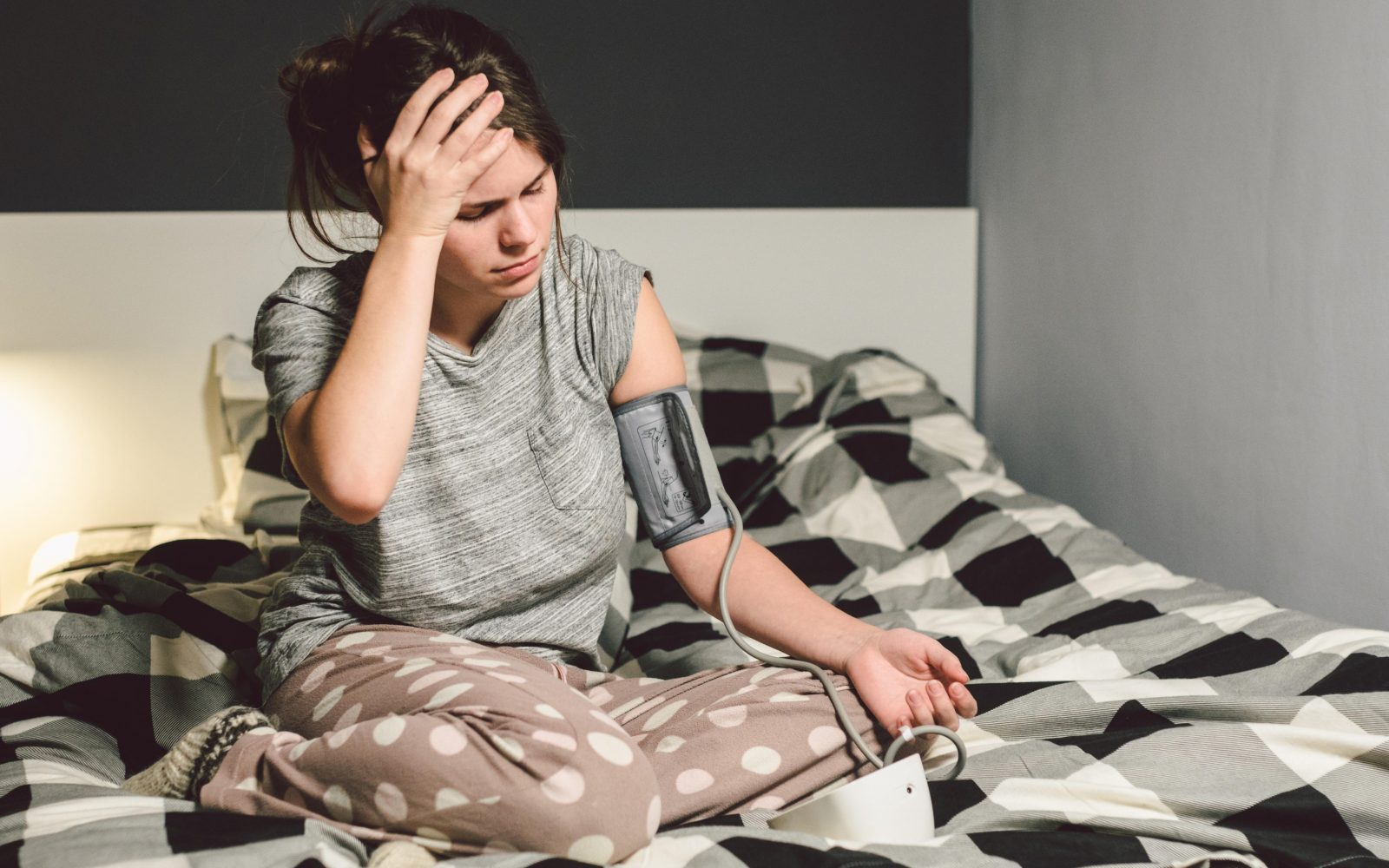 Mlada ženska v pižami sedi na postelji in si z merilnikom krvnega tlaka meri krvni tlak. Zaradi previsokega pritiska je zaskrbljena.