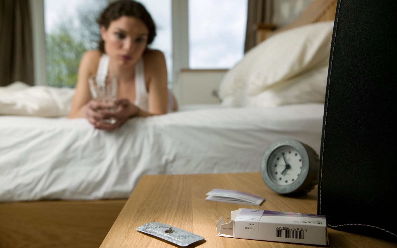 Prazen paket jutranjih tablet na nočni omarici z žensko, ki leži na postelji v ozadju