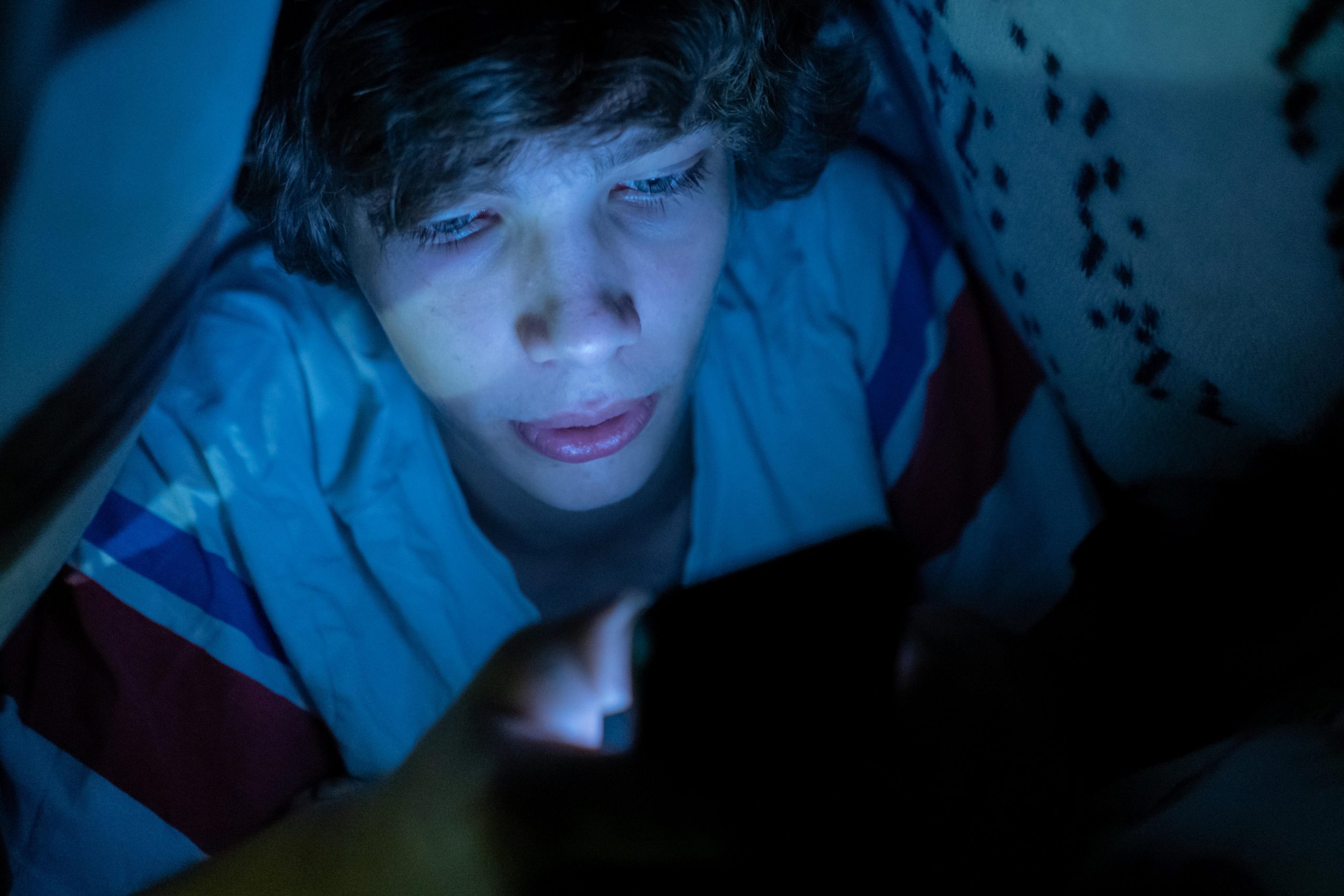 Najstnik na skrivaj pod odejo pasivno brska po telefonu, kar je znak prekomerne rabe zaslonov in ima dolgotrajne posledice na mentalno zdravje