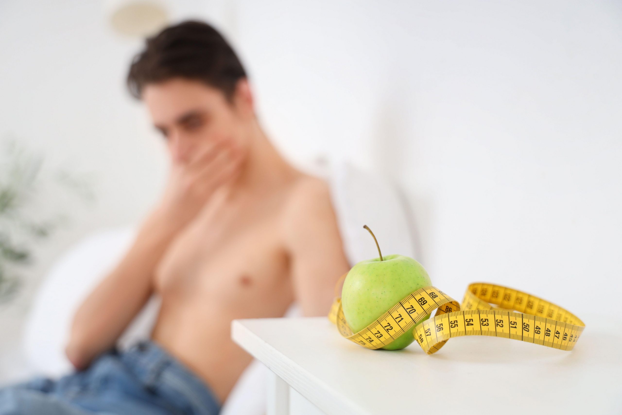 Moški sedi na postelji in zaskrbljeno gleda jabolko in meter, saj se sooča z motnjami hranjenja, ki so sicer pogostejše pri ženskah.