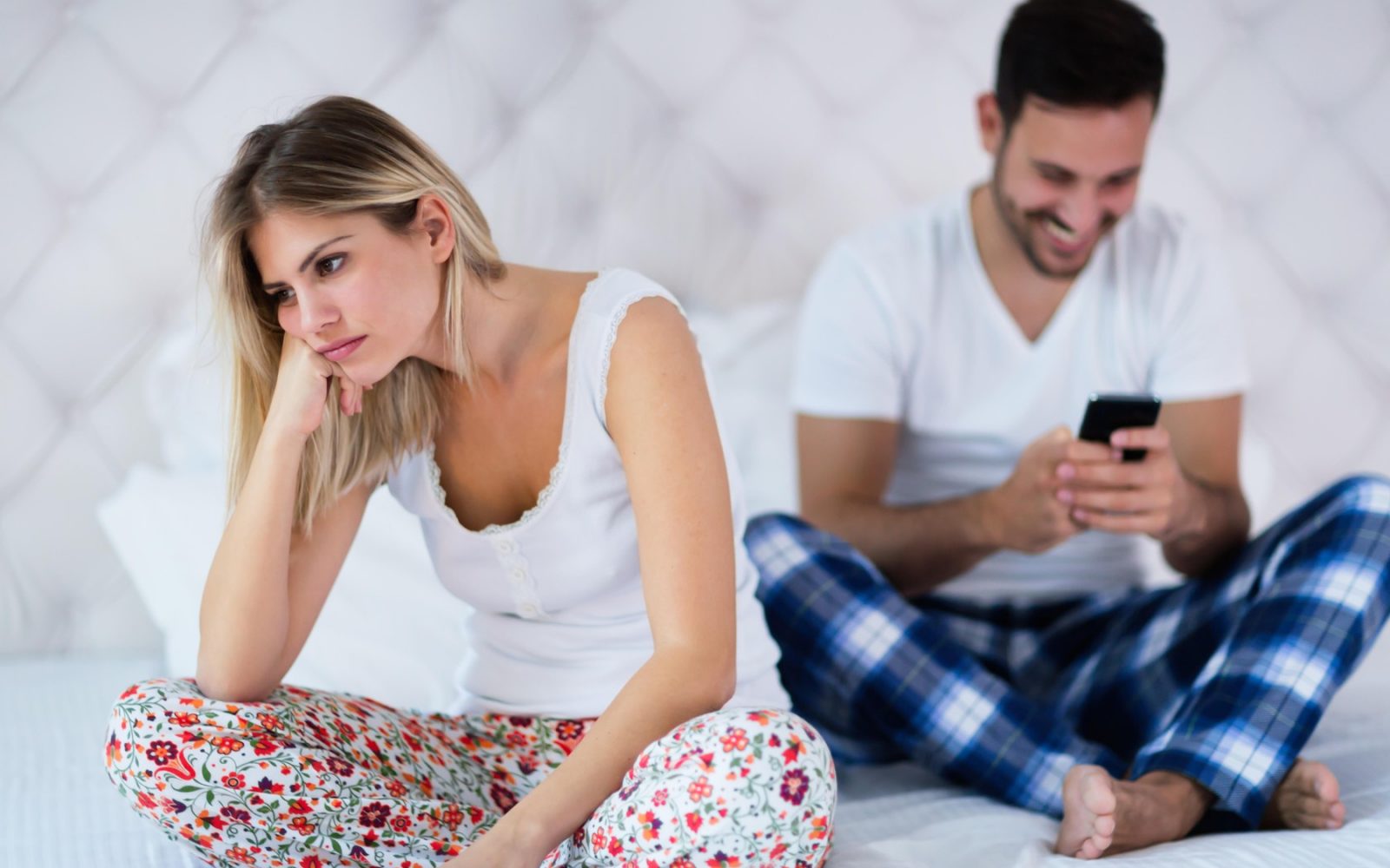Mlad par sedi na postelji. On tipka po telefonu, ona z nasmeškom je nejevoljno obrnjena stran.