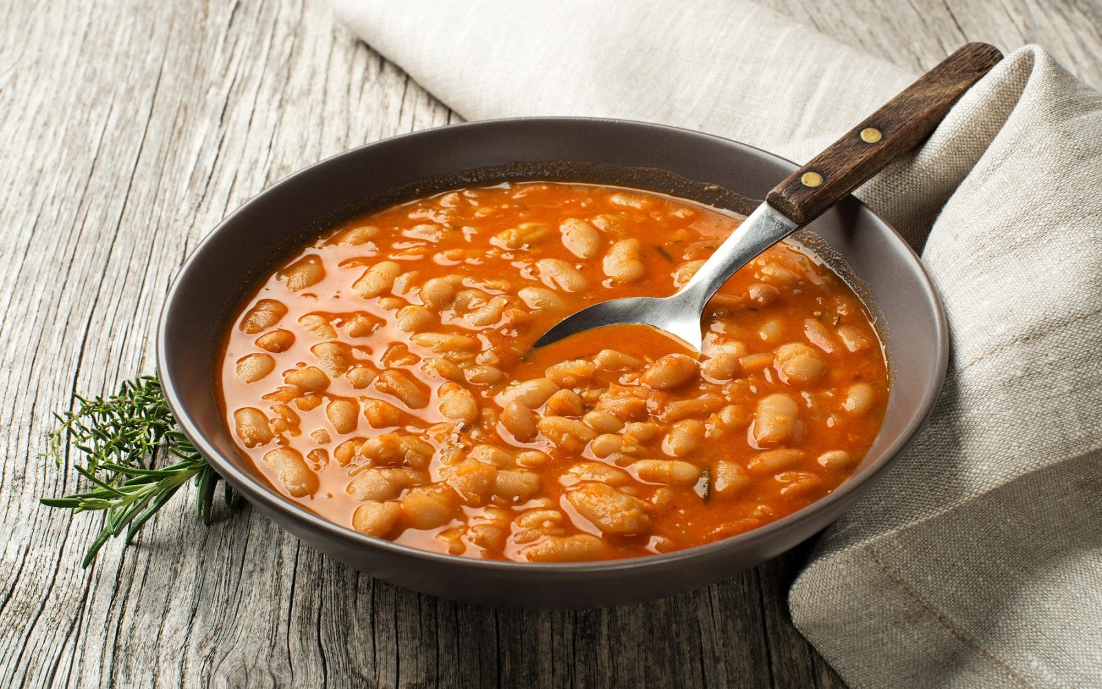 Domač pasulj z belim fižolom v paradižnikovi omaki – tradicionalna jed v globokem krožniku na lesenem ozadju.