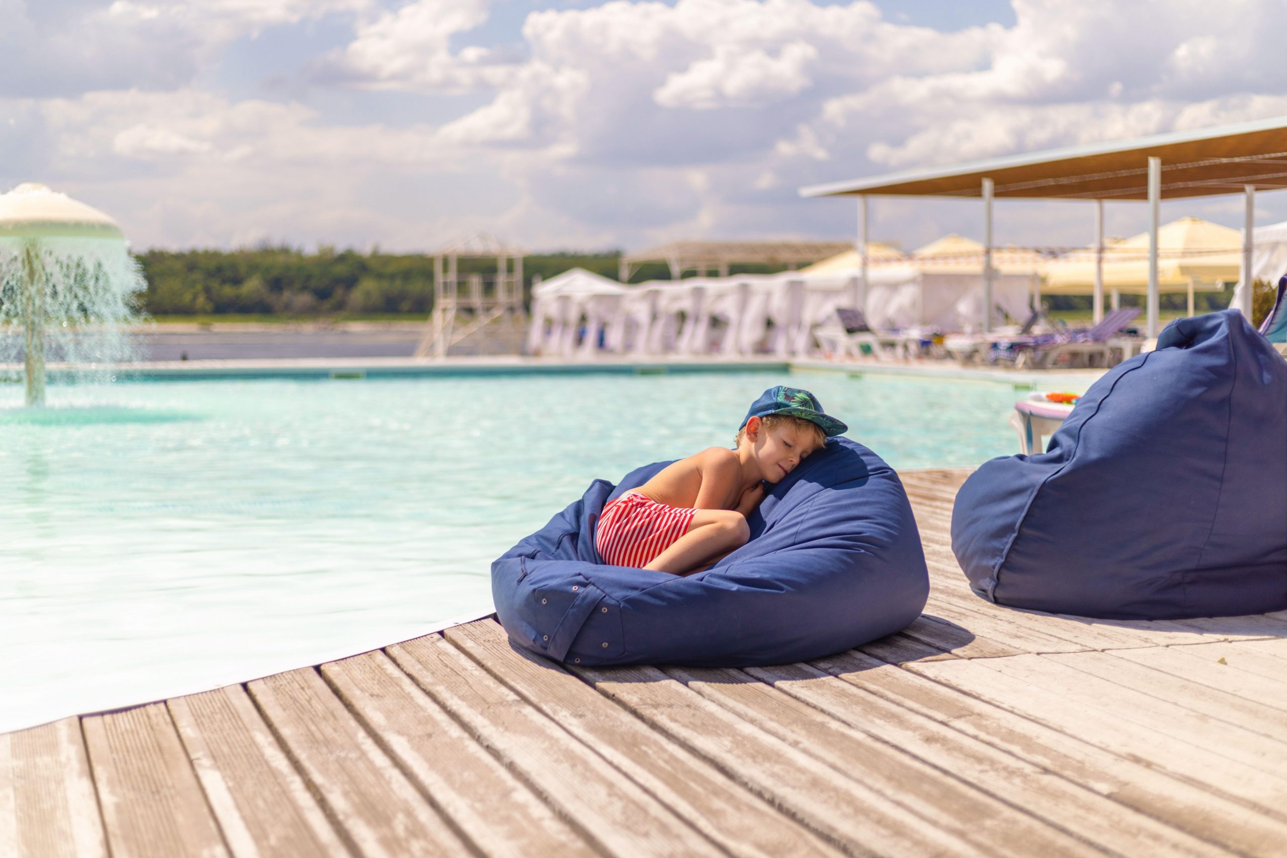 Otrok v črtastih hlačah in kapi počiva ob bazenu na blazini - počitek po jedi.