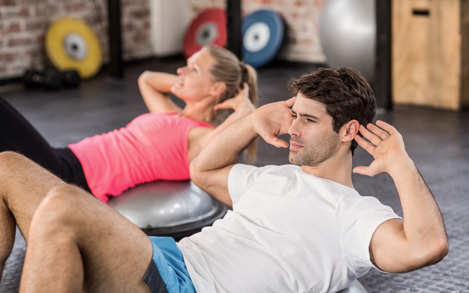Moški in ženska izvajata trebušnjake na polkrogli gimnastični žogi, s čimer krepita trebušne mišice in stabilnost trupa v fitnesu.