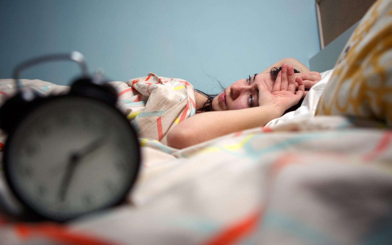 Ženska leži budna v postelji in zaskrbljeno gleda v budilko, ki kaže zgodnjo uro – simbol nespečnosti in težav s spanjem.