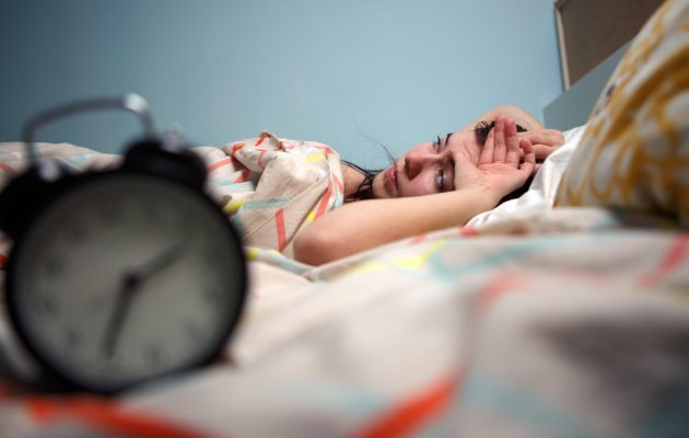 Ženska leži budna v postelji in zaskrbljeno gleda v budilko, ki kaže zgodnjo uro – simbol nespečnosti in težav s spanjem.