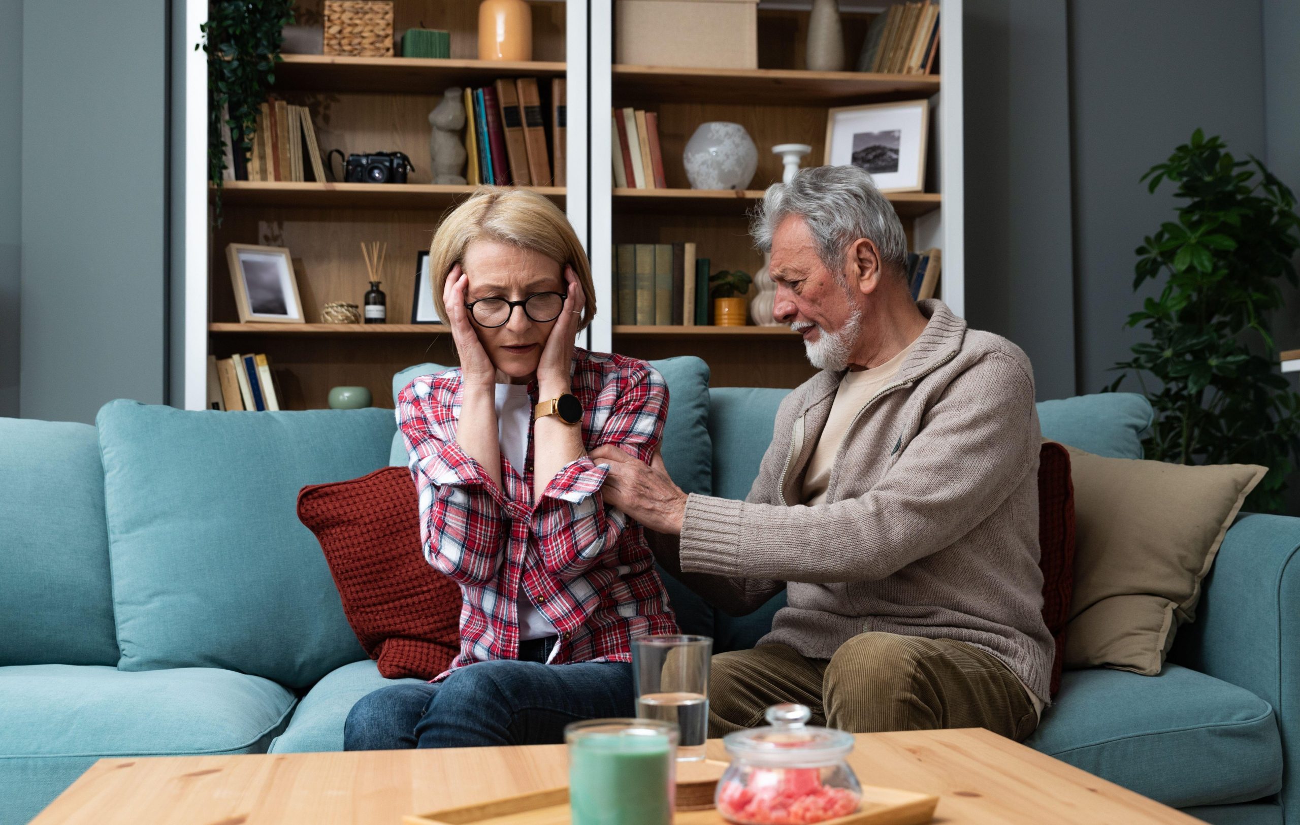 Starejši par sedi na kavču v dnevni sobi - starejši moški, skrbi za svojo bolno ženo, jo masira in objema, ker ima nenavadno redko bolezen.