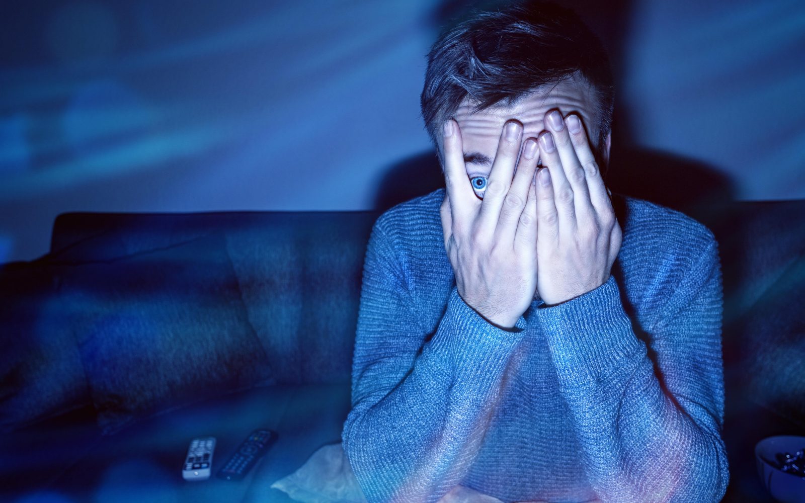 Moški si zakriva oči ob gledanju grozljivke, ki je stimulacija za možgane in ni priporočljiva pred spanjem.