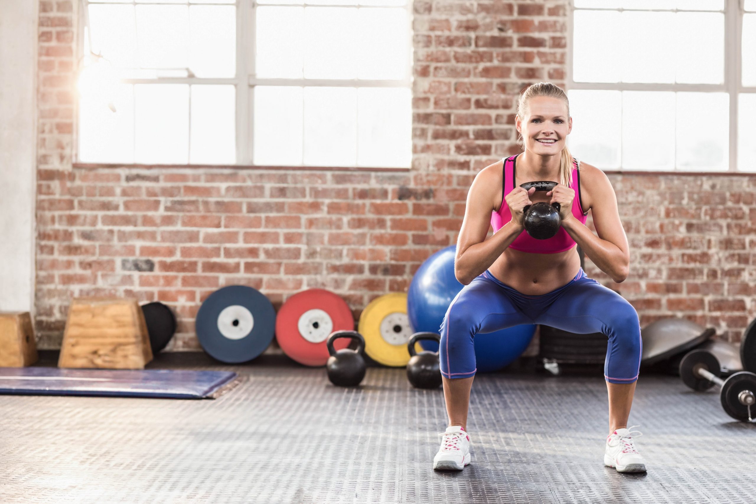 Ženska v fitnesu, polnem pisanih uteži, dela počep in pri tem drži kettlebell utež pred prsmi.