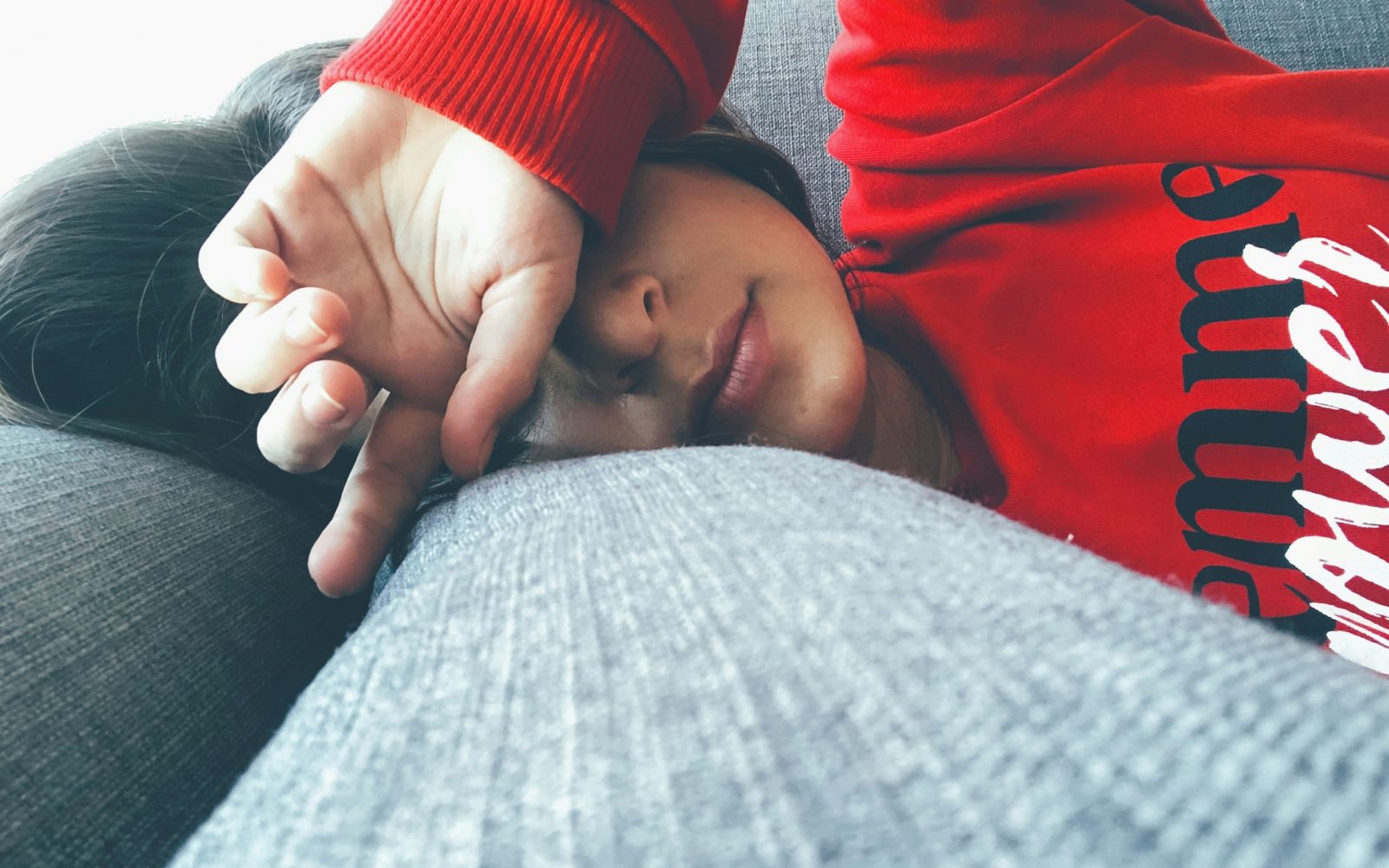 Utrujena ženska v rdečem puloverju leži na kavču, z roko si pokriva oči