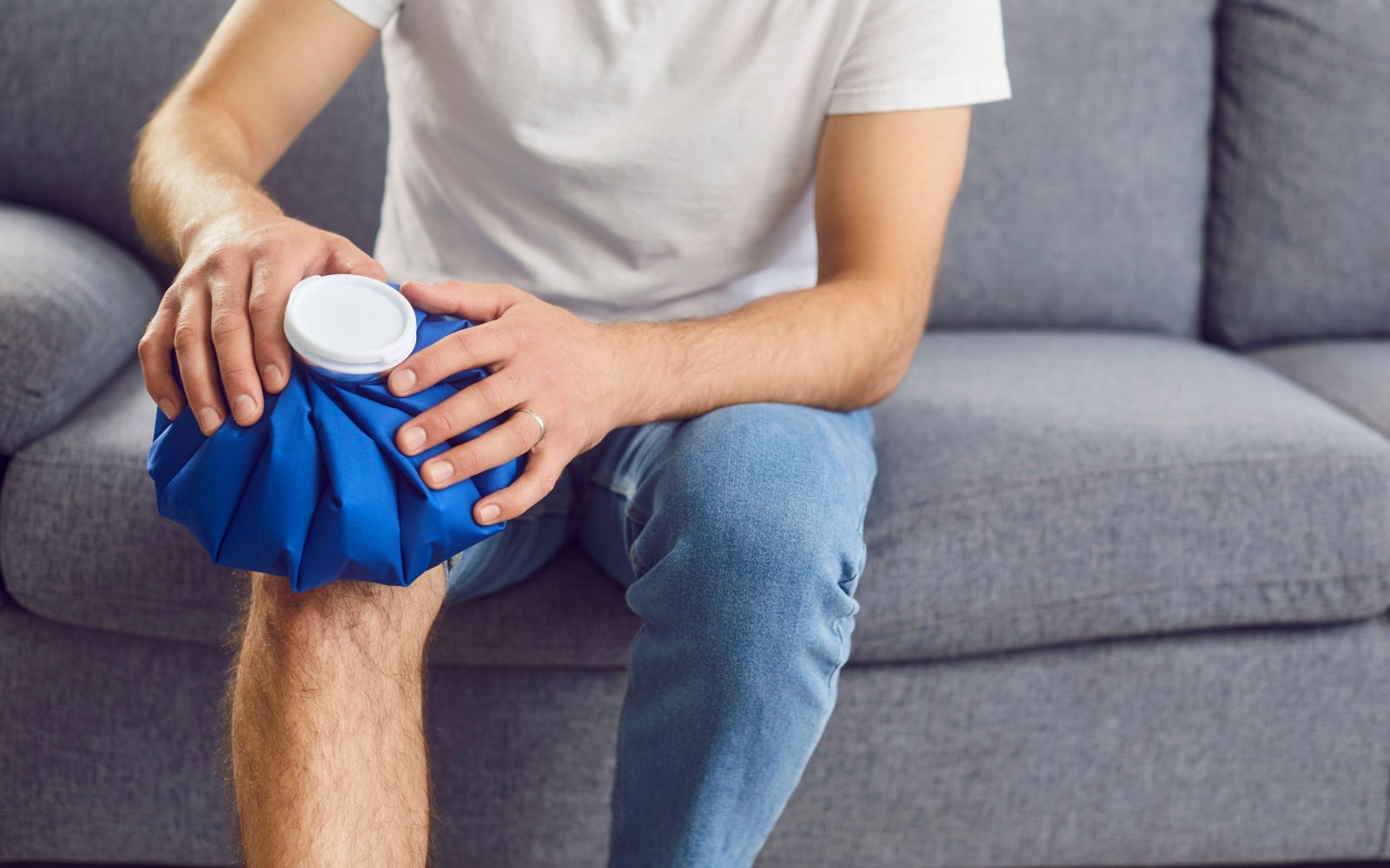 Mlad moški z artritisom sedi na kavču doma in drži leden obkladek na poškodovanem kolenu, da zmanjša bolečino in oteklino.