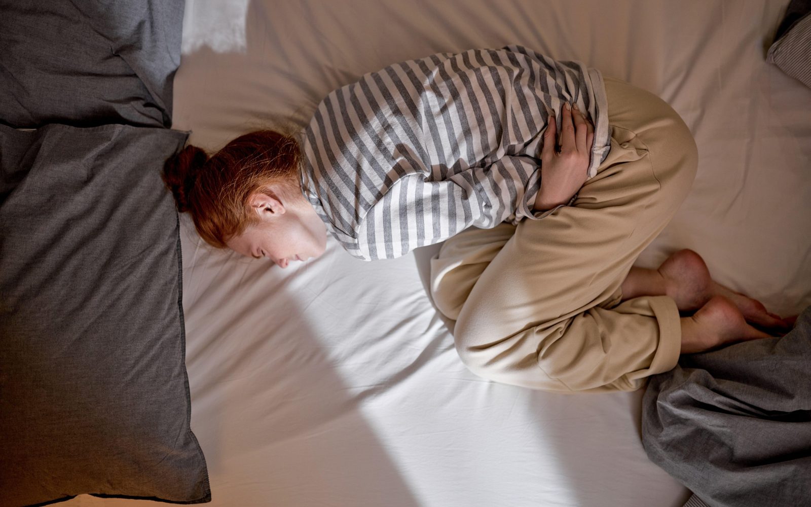 Mlada rdečelasa ženska leži v postelji, se drži za trebuh in se zvija od bolečin, ki jih povzročajo menstrualni krči.