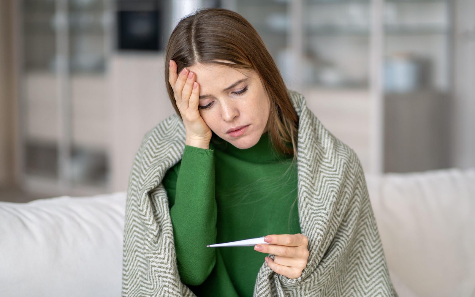 Utrujena prehlajena ženska sedi na kavču zavita v deko, se drži za bolečo glavo in gleda termometer - jesen sezona prehladov