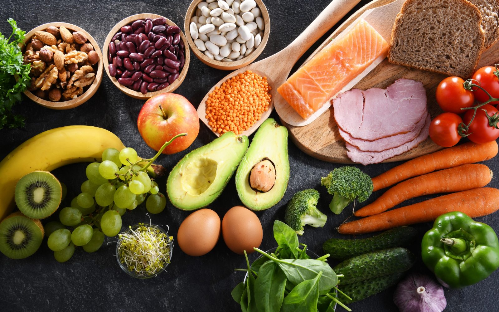 Uravnotežena in raznolika prehrana - na mizi so hranljiva živila, kot je kruh, losos, paradižnik, korenje, brokoli, kumara, bazilika, paprika, česen, jajca, kalčlki, avokado, leča, fižol, oreščki, banana, kivi, grozdje, peteršilj, jabolko, meso.