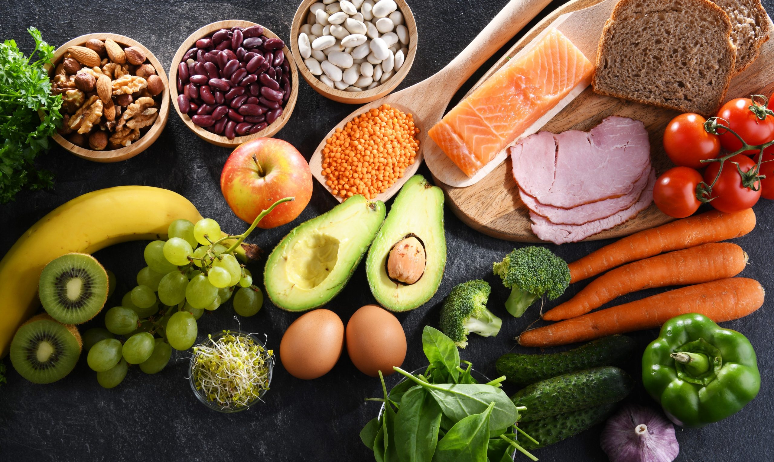 Uravnotežena in raznolika prehrana - na mizi so hranljiva živila, kot je kruh, losos, paradižnik, korenje, brokoli, kumara, bazilika, paprika, česen, jajca, kalčlki, avokado, leča, fižol, oreščki, banana, kivi, grozdje, peteršilj, jabolko, meso.