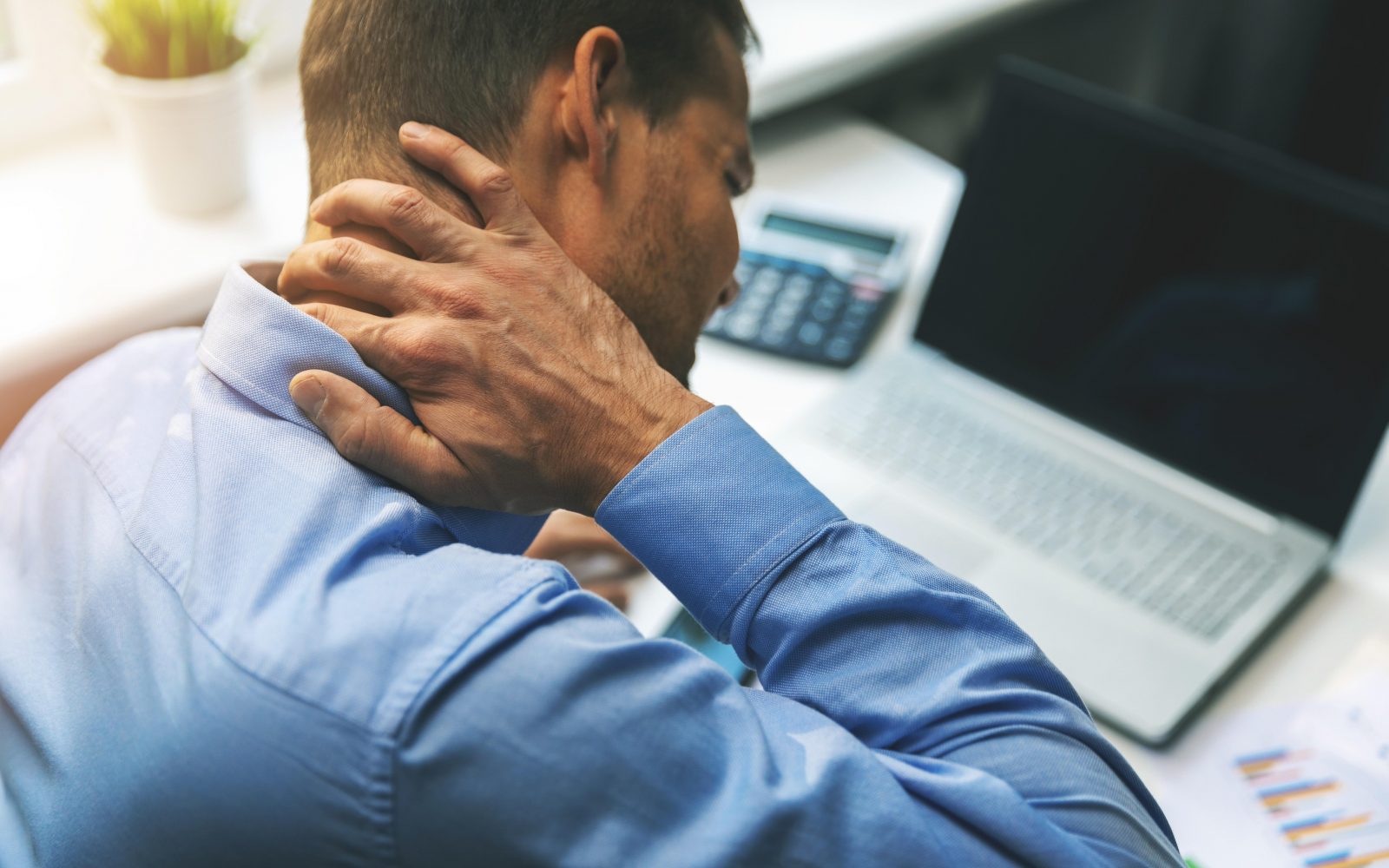Moški sedi za mizo za računalnikom in se drži za vrat zaradi bolečine, ki jo povzroča nepravilna drža in sedeč življenjski slog