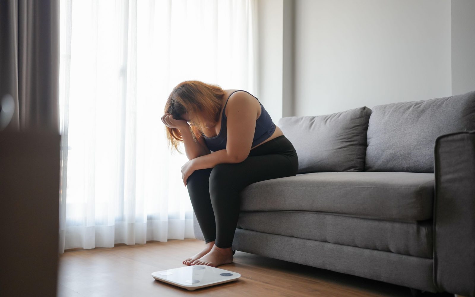 Debelejša ženska sedi na kavču, obupana zaradi neuspešnih diet in hujšanja, na tleh je tehtnica.