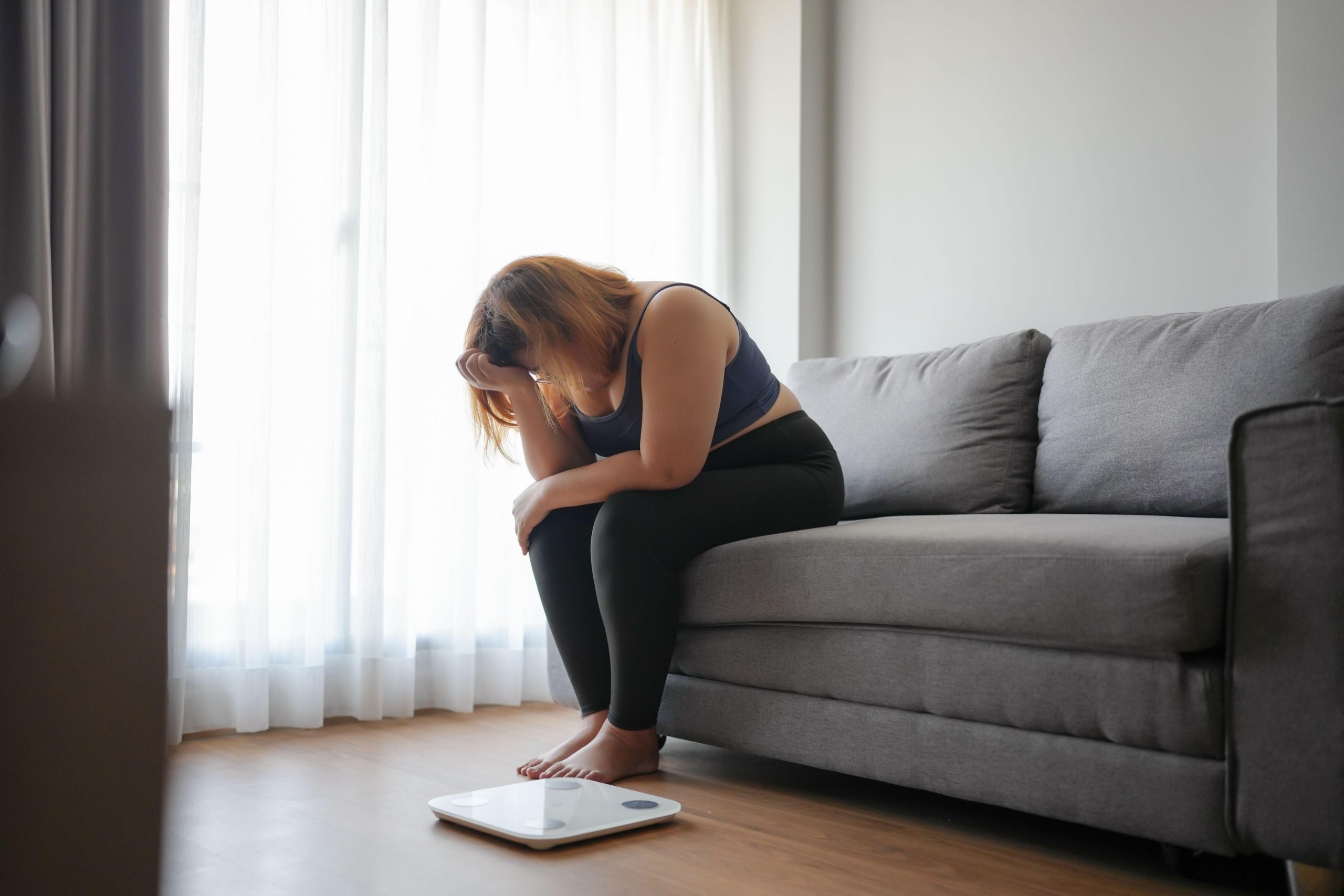 Debelejša ženska sedi na kavču, obupana zaradi neuspešnih diet in hujšanja, na tleh je tehtnica.